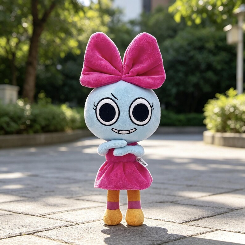May include: A plush toy featuring a light blue round head with large black eyes, a small grin, and a large bright pink bow on top. The figure wears a bright pink dress, orange legs with pink bands, and yellow feet, standing upright on a paved surface.