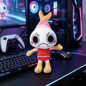 May include: A plush toy featuring a white, round head with large black eyes and a frowning mouth, topped with a pink shrimp-like tail. The character wears a red shirt with pink sleeves and tan-colored hands and feet, standing upright.