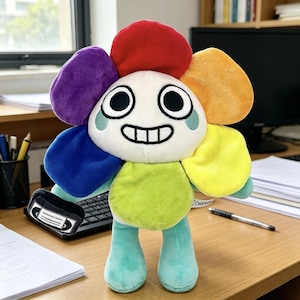 Op de afbeelding: Een pluche speelgoedfiguur in de vorm van een bloem met een wit gezicht, grote zwarte ogen en een brede glimlach. De bloemblaadjes zijn paars, rood, oranje, geel en groen. Het lichaam en de ledematen zijn lichtgroen en één hand houdt een zwarte cassette.