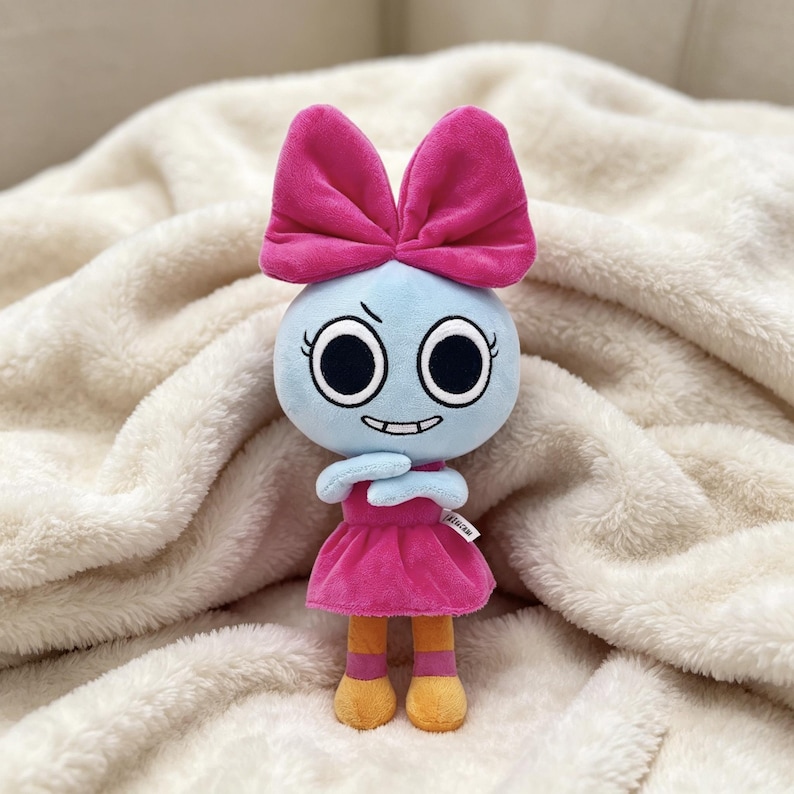 May include: A plush doll with a light blue round head, large black eyes, and a mischievous expression. It wears a large bright pink bow on its head, a matching pink dress, and has yellow legs with pink bands. A small white tag is visible on the side.