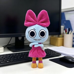 May include: A plush toy featuring a light blue round head with large black eyes, a small grin, and a prominent bright pink bow on top. The figure wears a matching pink dress and has orange legs with pink accents at the ankles and feet.