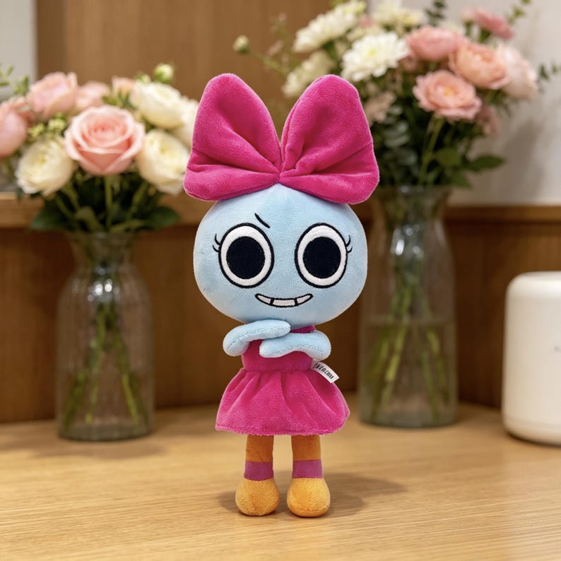 May include: A plush doll with a light blue round head, large black eyes, and a mischievous grin. It features a large bright pink bow on top of its head, a matching pink dress, and orange legs with pink accents. A small white tag is visible on the side.