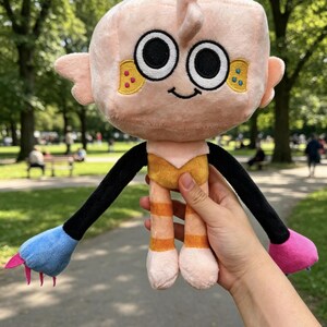 May include: A plush toy with a large, square, light pink head, black eyes, and a small smile. It features long black arms ending in one blue and one pink hand with small claws. The body is yellow and orange, with orange striped legs.