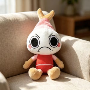 May include: A plush toy featuring a white, round head with large black eyes, a frowning mouth, and pink circular cheeks. The top of the head has a tan, shrimp-like tail fin. The body is dressed in a red shirt with light pink sleeves and tan limbs.