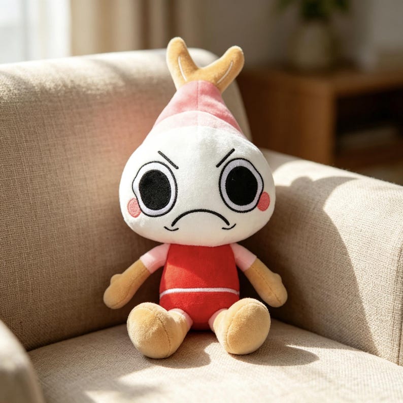 May include: A plush toy featuring a white, round head with large black eyes, a frowning mouth, and pink circular cheeks. The top of the head has a tan, shrimp-like tail fin. The body is dressed in a red shirt with light pink sleeves and tan limbs.