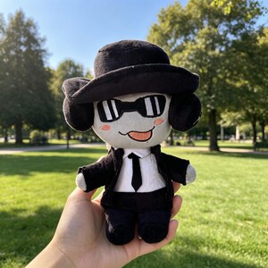 May include: A plush doll wearing a black fedora, black sunglasses, a black suit jacket, and a black tie over a white shirt. The doll has a light gray face with pink blush on the cheeks and a small orange tongue sticking out from its mouth.