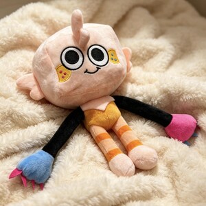 May include: A plush toy featuring a light peach-colored square head with large black and white eyes, a small smile, and yellow cheek patches. The body has black arms, orange striped legs, and mismatched hands in blue and pink with small claws.