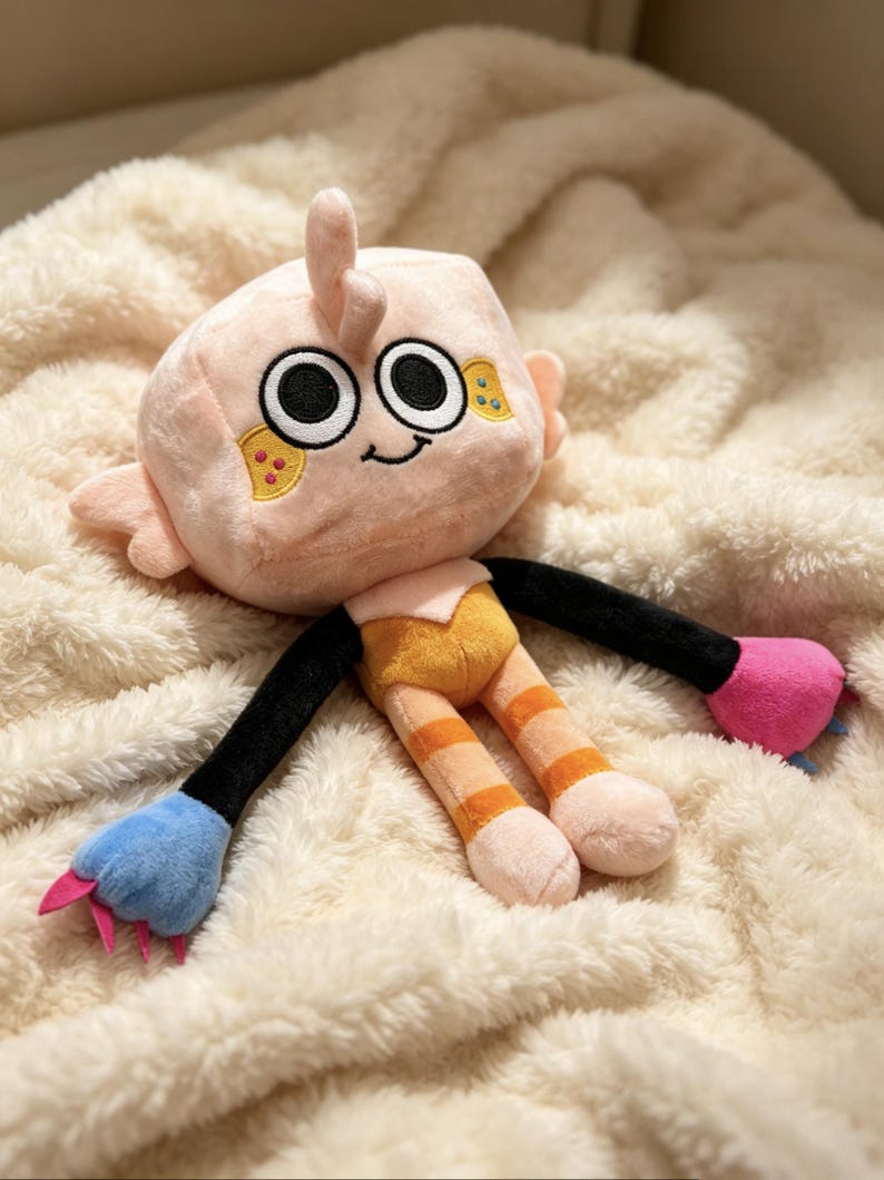 May include: A plush toy featuring a light peach-colored square head with large black and white eyes, a small smile, and yellow cheek patches. The body has black arms, orange striped legs, and mismatched hands in blue and pink with small claws.