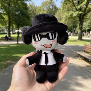 May include: A plush doll featuring a light gray face with pink cheeks and a tongue sticking out. The doll wears a black fedora, black sunglasses, a black suit jacket, a white shirt, and a black necktie. The plush has black ears and limbs.
