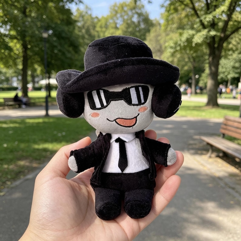 May include: A plush doll featuring a light gray face with pink cheeks and a tongue sticking out. The doll wears a black fedora, black sunglasses, a black suit jacket, a white shirt, and a black necktie. The plush has black ears and limbs.