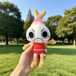 May include: A plush toy featuring a white, round head with a pointed pink top, large black eyes, and an embroidered frowning mouth. The character has a red torso, pink limbs, and tan-colored hands and feet, with a small tan tail-like protrusion.