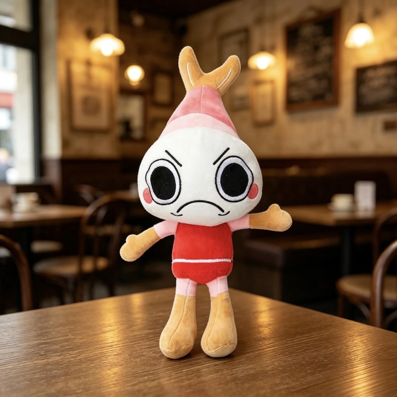 May include: A plush toy shaped like a shrimp with a white head, large black eyes, and a frowning mouth. It features a pink pointed top, a red torso, and tan-colored arms and legs. The character has a small tail-like structure on its head.
