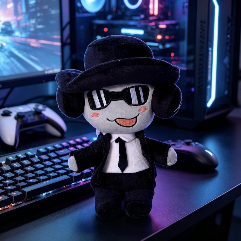 May include: A plush doll featuring a light gray head with embroidered black sunglasses, pink cheeks, and a tongue sticking out. The doll wears a black fedora, a black suit jacket, a white shirt, and a black tie, with black plush hands and feet.