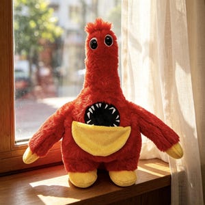 Op de afbeelding: Een knuffel in de vorm van een lang, pluizig rood wezen met een lange nek, twee grote zwarte ogen en een brede, ronde mond vol scherpe witte tanden. Het wezen heeft gele handen en voeten en een gele buidel op het middenstuk.
