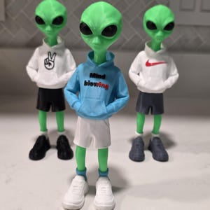 Könnte beinhalten: Drei grüne Alien-Figuren in aufrechter Position. Die mittlere Figur trägt einen hellblauen Kapuzenpullover mit dem Text "Mind blowing" in Schwarz und Rot, weiße Shorts und weiße Sneaker. Die beiden äußeren Figuren tragen weiße Kapuzenpullover und schwarze Shorts.