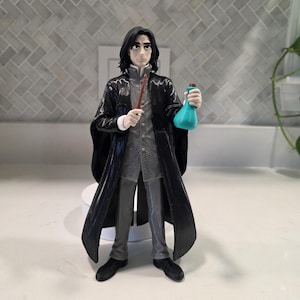 May include: A figurine of a person with shoulder-length black hair, wearing a long black coat over a grey collared shirt and grey trousers. The figure holds a thin brown wand in the right hand and a small, round, teal-colored potion bottle in the left.