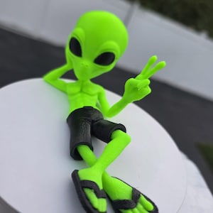 Könnte beinhalten: Eine leuchtend neongrüne Alien-Figur mit großen schwarzen ovalen Augen, die eine schwarze Shorts und schwarze Flip-Flops trägt. Das Alien befindet sich in einer liegenden Pose, stützt den Kopf auf eine Hand und zeigt mit der anderen das Friedenszeichen.