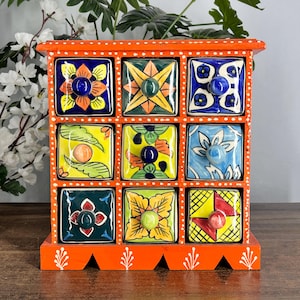 May include: A wooden storage chest with nine square drawers, each featuring a unique hand-painted floral or geometric pattern in vibrant colours. The frame is painted bright orange with white polka dots and white leaf motifs at the base.