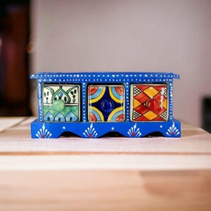 May include: A blue wooden storage chest featuring three square drawers with hand-painted ceramic fronts. The left drawer has green floral patterns, the centre has blue and yellow geometric shapes, and the right has a red and orange diamond grid.