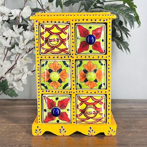 May include: A yellow wooden storage chest with six square drawers. Each drawer front features hand-painted ceramic tiles with colorful floral and geometric patterns in red, orange, green, and yellow. The frame has blue dot accents.