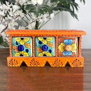 Può includere: Un mobiletto rettangolare in legno arancione con tre cassetti. I due cassetti a sinistra presentano motivi floreali gialli e arancioni su sfondo turchese con pomelli blu. Il cassetto a destra ha un fiore blu e giallo su sfondo marrone.