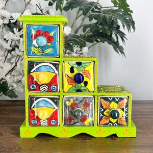 May include: A lime green wooden tiered storage unit with six square drawers. Each drawer features a unique hand-painted ceramic front with colorful floral, geometric, and abstract patterns, each equipped with a small round knob for opening.