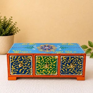 Pode incluir: Um organizador de secretária retangular de madeira com três gavetas e molduras cor de laranja. A superfície superior é pintada de azul claro com um motivo floral central. As duas gavetas exteriores são azul-escuras com vinhas amarelas.