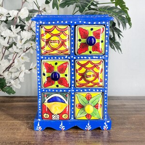 May include: A blue wooden storage chest featuring six square drawers with hand-painted ceramic fronts. Each drawer displays vibrant, multi-colored floral and geometric patterns in red, yellow, green, and blue, finished with small round knobs.