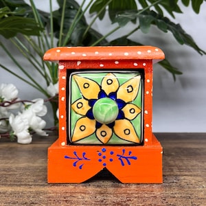 Könnte beinhalten: Eine kleine orangefarbene Holztruhe mit einer einzelnen Schublade, deren Front mit einer handbemalten Keramikfliese verziert ist. Die Fliese zeigt ein gelb-blaues Blumenmuster auf hellgrünem Grund. Der orange Rahmen hat weiße Punkte und blaue Akzente.