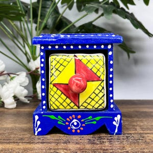 May include: A small blue wooden storage chest with a single drawer featuring a yellow and red geometric pattern. The blue frame is decorated with white dots and green and orange floral accents, and the drawer has a round red knob.