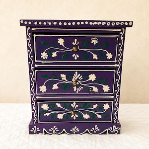 May include: A small purple wooden chest with three drawers, each featuring hand-painted white floral vines and green leaf accents. The chest has a scalloped base and a top edge decorated with a row of white dots, finished with small brass knobs.