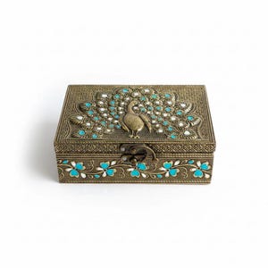 May include: A rectangular antique brass-toned metal box featuring an embossed peacock design on the lid. The peacock is surrounded by intricate patterns accented with small turquoise and white enamel inlays. A metal latch secures the lid.