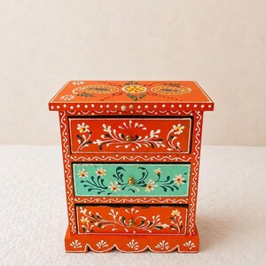Könnte beinhalten: Eine kleine Holzkommode mit leuchtend orangefarbenem Anstrich und handgemalten weißen Blumenmustern. Das Möbelstück verfügt über drei Schubladen, wobei die mittlere in einem kontrastierenden Türkis gehalten ist und jede einen kleinen Messingknopf besitzt.