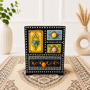 May include: A black wooden chest of drawers featuring three small upper drawers and one large lower drawer. The drawers are painted in yellow, light blue, and green with colorful floral patterns and bright round knobs in blue, yellow, pink, and orange.