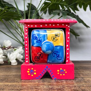 May include: A small red wooden storage box with white polka dots and a central square ceramic drawer. The drawer face is divided into four quadrants of blue, yellow, red, and blue, each featuring a hand-painted black floral line drawing.