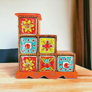Könnte beinhalten: Ein gestuftes orangefarbenes Holzregal mit sechs Schubladen, die jeweils mit handgemalten Blumen- und geometrischen Mustern in leuchtendem Rot, Gelb und Türkis verziert sind. Die Struktur hat ein stufenförmiges Design mit gewelltem Rand.