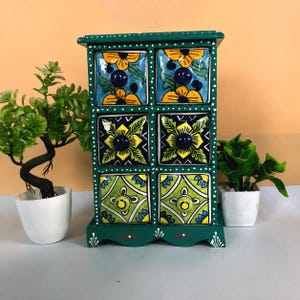May include: A dark green wooden storage chest featuring six drawers with hand-painted ceramic fronts. Each drawer displays distinct floral and geometric patterns in shades of yellow, blue, and green, accented by small dark blue or yellow knobs.