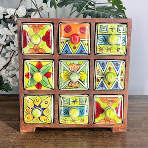 May include: A wooden storage chest featuring nine square drawers with hand-painted ceramic fronts. Each drawer displays a unique colorful pattern including floral, geometric, and abstract designs in shades of orange, green, blue, and yellow.