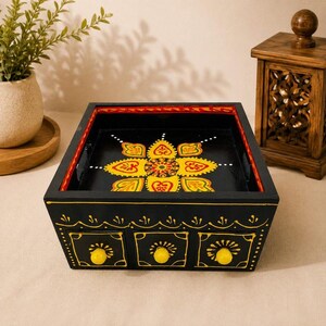 May include: A black wooden desktop organizer featuring three small drawers with yellow knobs. The front and top surfaces are decorated with hand-painted yellow, red, and green floral patterns. The box has a square shape with a flat open top tray.
