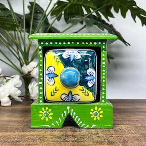 Op de afbeelding: Een kleine groene houten opbergdoos met één lade met een vierkant keramisch front. De ladefront toont een geel en donkergroen bloemmotief met een centrale blauwe knop. Het groene frame is versierd met witte stippen en gele bloemen.