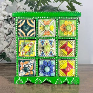 May include: A bright green wooden storage chest featuring nine square drawers, each with a unique hand-painted ceramic front. The drawers display colorful floral, geometric, and grid patterns in shades of yellow, orange, blue, and red, with small round knobs.