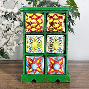 May include: A green wooden storage chest with six square drawers. The top two drawers feature a yellow and red scale pattern, the middle two have a green, blue, and white geometric design, and the bottom two display a red and yellow floral motif.