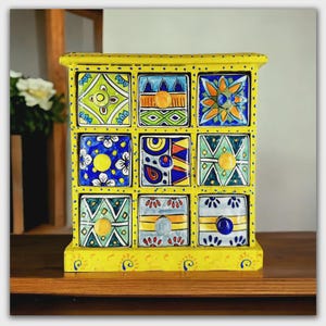 May include: A yellow wooden storage chest featuring nine square drawers, each decorated with unique hand-painted geometric and floral patterns in blue, green, orange, and white. Each drawer is equipped with a small, solid-colored round knob.