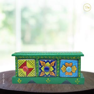 Può includere: Un bauletto in legno verde con tre cassetti quadrati dotati di frontali in ceramica dipinti a mano. Il cassetto sinistro presenta una griglia gialla con triangoli rossi, quello centrale un motivo floreale blu e quello destro un fiore giallo.