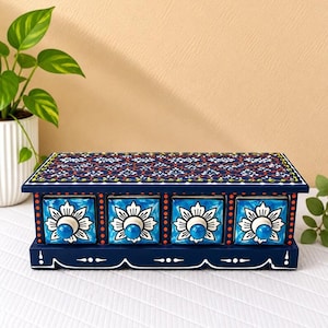 Puede incluir: Un cofre de almacenamiento rectangular de madera con cuatro cajones con patrones florales en blanco y azul. El cofre está pintado de azul oscuro con detalles de puntos naranjas y un motivo floral detallado en la parte superior.