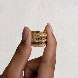 May include: A stack of six gold-toned rings featuring various designs, including bands with clear white stones, olive green stones, and baguette-cut stones. The rings are arranged vertically, showcasing different textures and stone settings in a metallic finish.
