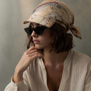 May include: A patterned headscarf with a visor, paired with black sunglasses and a cream-colored blouse. The headscarf features a patchwork design in muted tones. The sunglasses have a cat-eye shape.