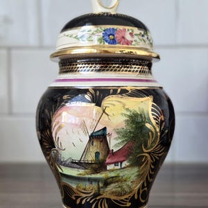 Puede incluir: Un jarrón de cerámica negro y dorado con tapa, que presenta una escena de paisaje pintada a mano con un molino de viento y una casa. La tapa está adornada con motivos florales y un asa decorativa.