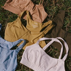May include: An assortment of women's sports bras in various colors, including light blue, mustard yellow, olive green, tan, and off-white. The bras are arranged on a bed of green grass, showcasing their different designs and styles.