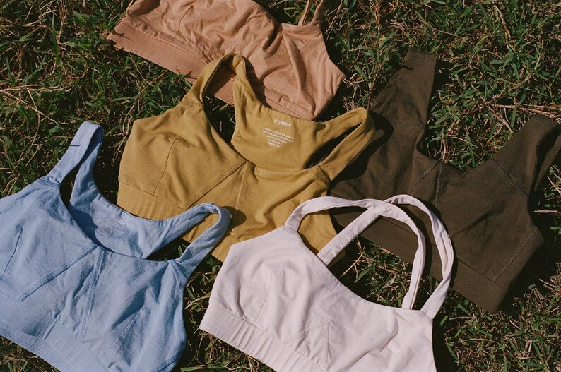 May include: An assortment of women's sports bras in various colors, including light blue, mustard yellow, olive green, tan, and off-white. The bras are arranged on a bed of green grass, showcasing their different designs and styles.